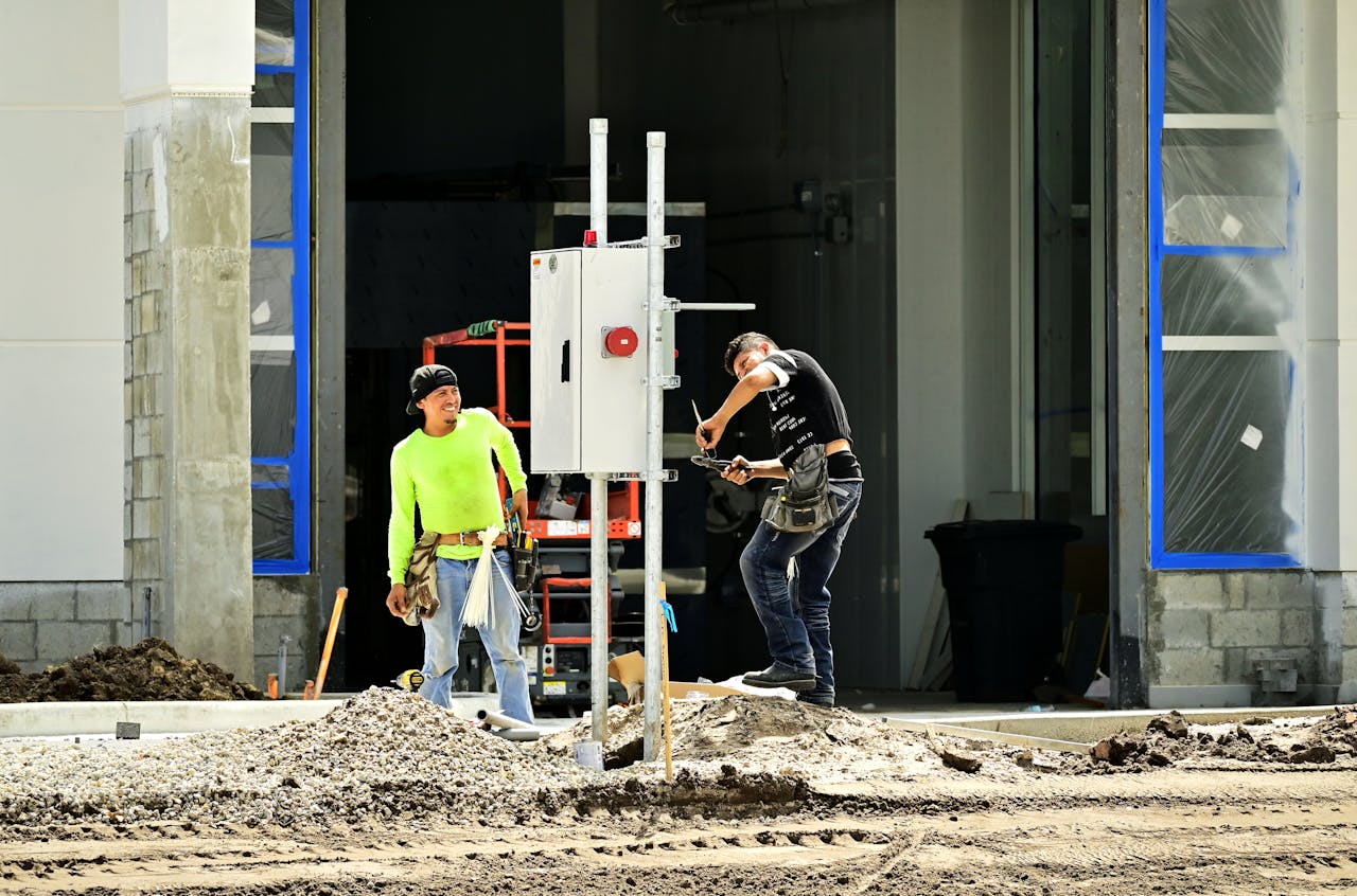 Two construction workers install an electrical panel outside a building site, wearing safety gear.